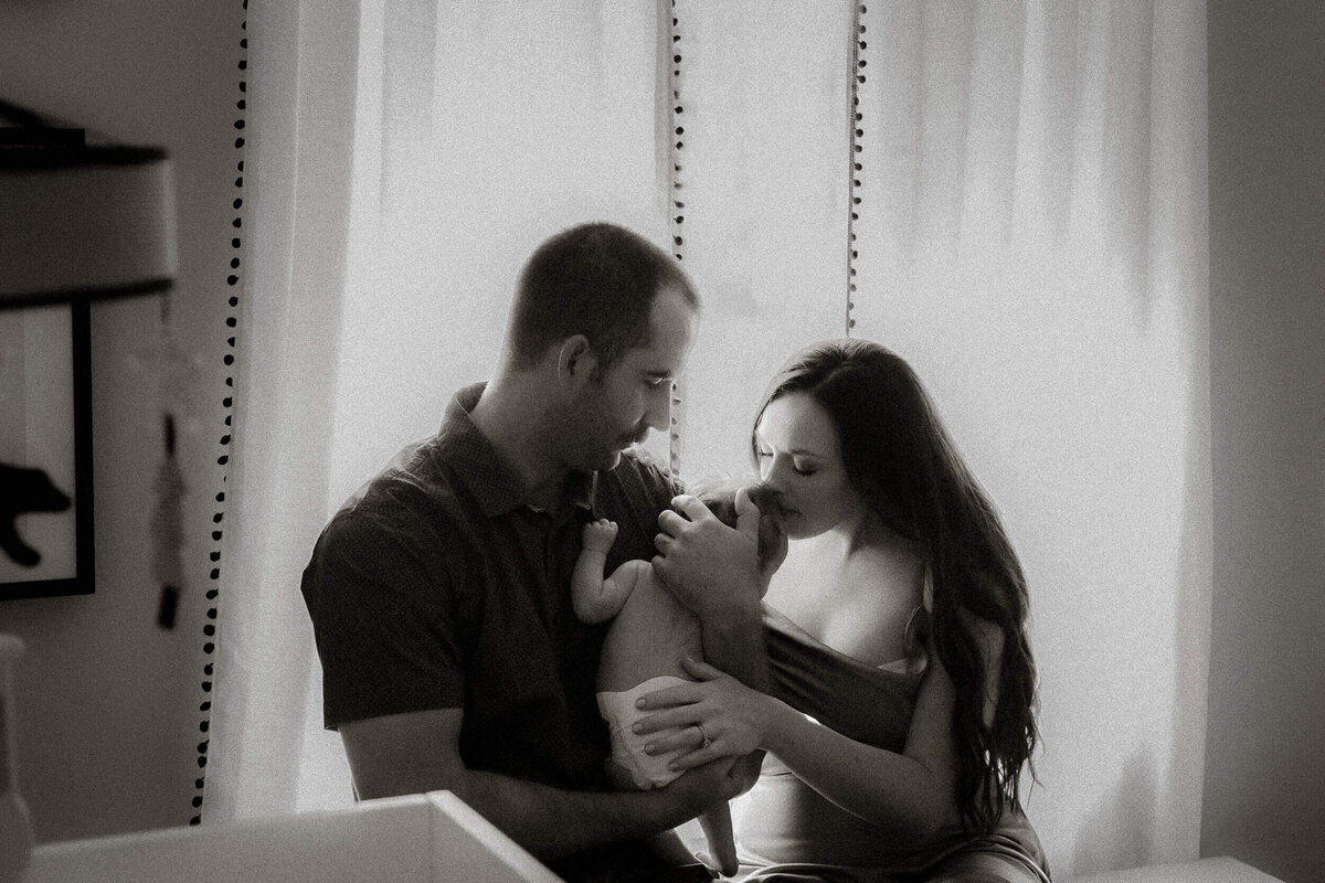 A family of three sitting by the window with dad holding their newborn while mom gently kisses him on the head in black and white.