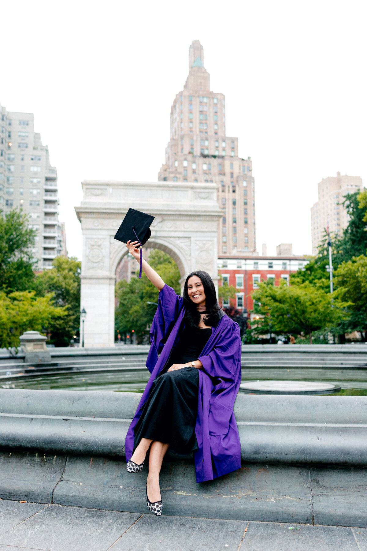 NYU MBA graduate smiling while holding cap at the fountain in Washington Square Park.
