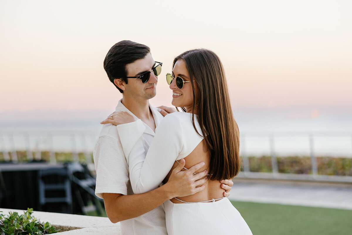 The engaged couple poses confidently in front of the scenic ocean view, both wearing stylish sunglasses, capturing a chic and modern engagement session at Marriott Hotel Fort Lauderdale.