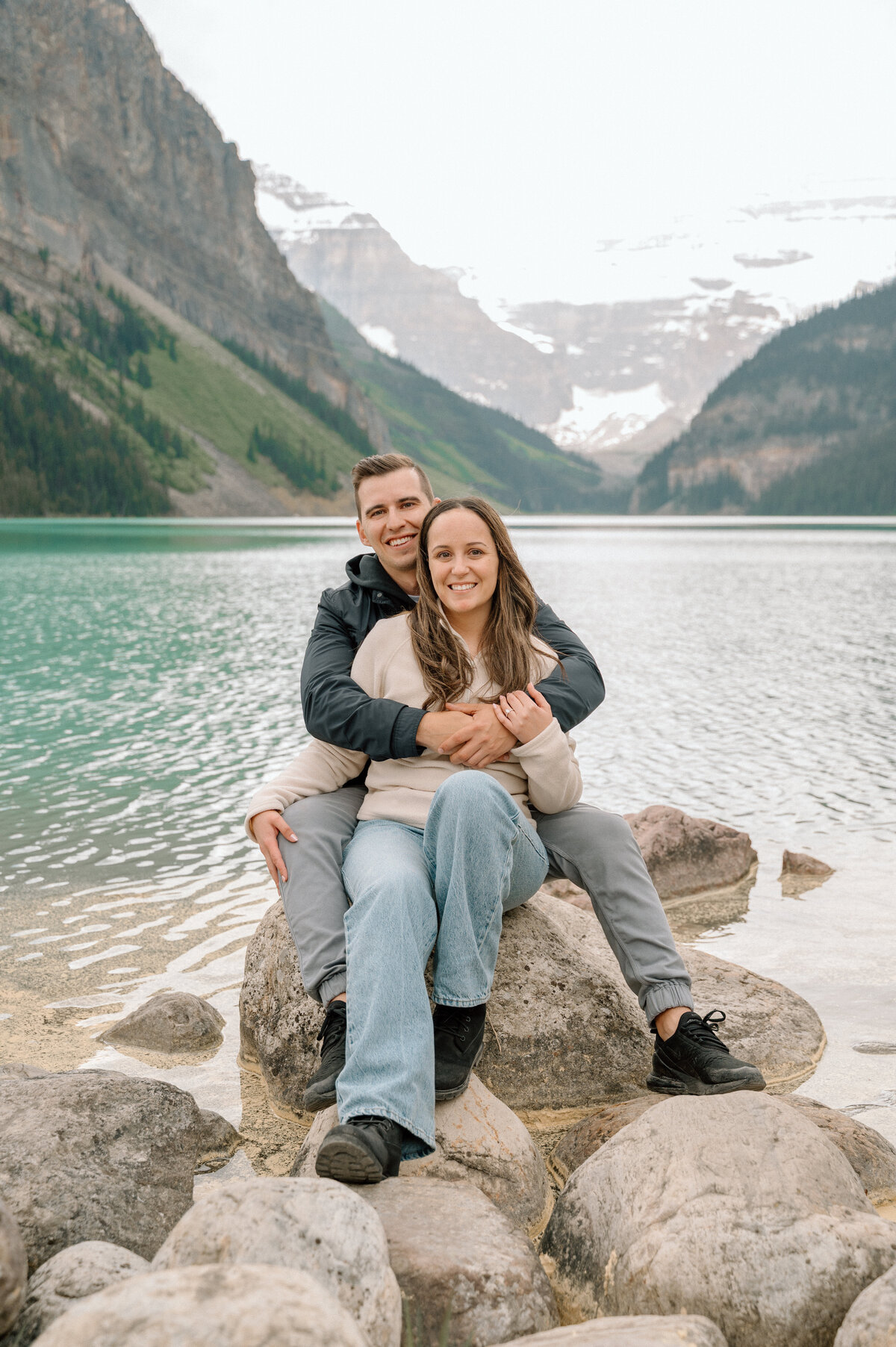 Engagement photo with couple seated on large rock by Lake Louise shoreline.