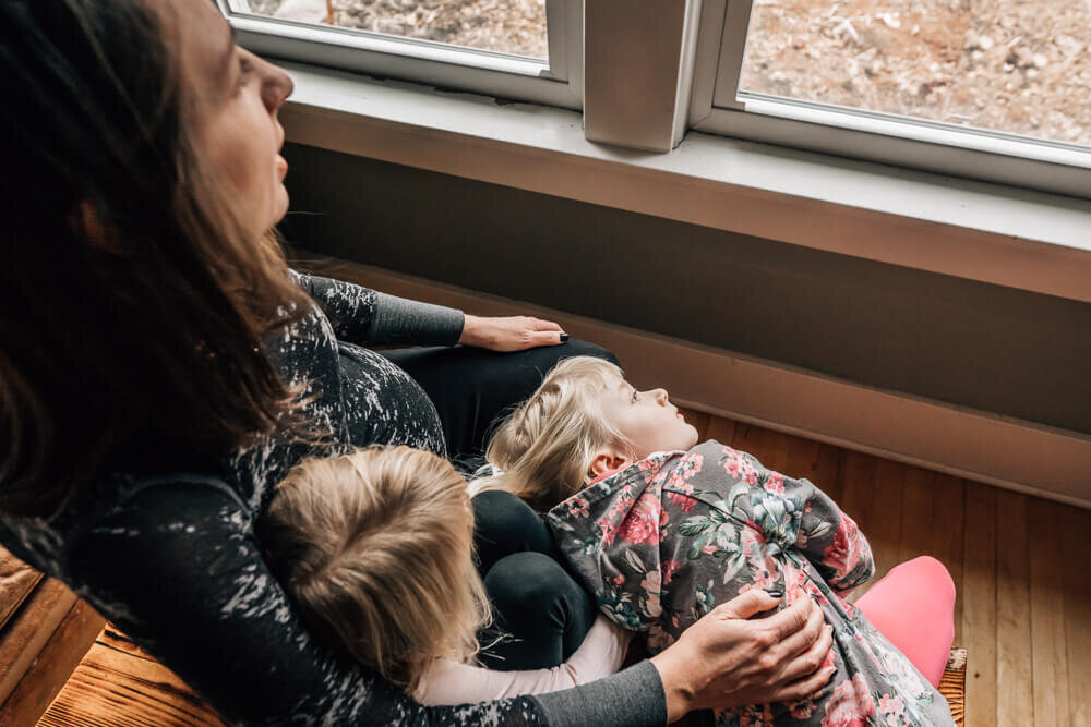 A pregnant mother sits with her two young girls, all gazing out the window, during an in home maternity photography session by Kate Simpson.