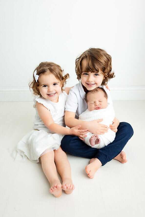 Siblings hold their new baby brother in a white studio.