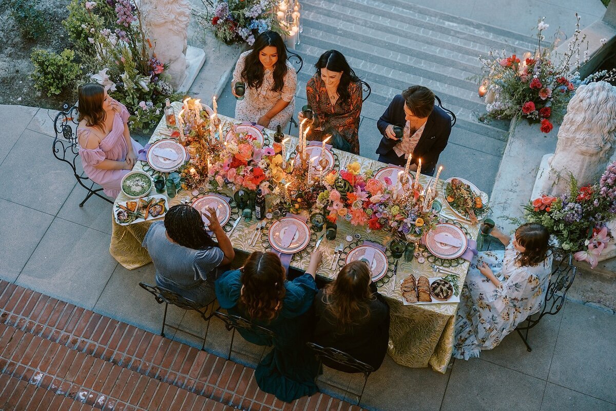 Wrensmoor Castle maximalist alfresco dinner party reception  surrounded by candelabras and colorful spring garden-flowers. At sunset guests are seated in wrought iron bistro chairs.