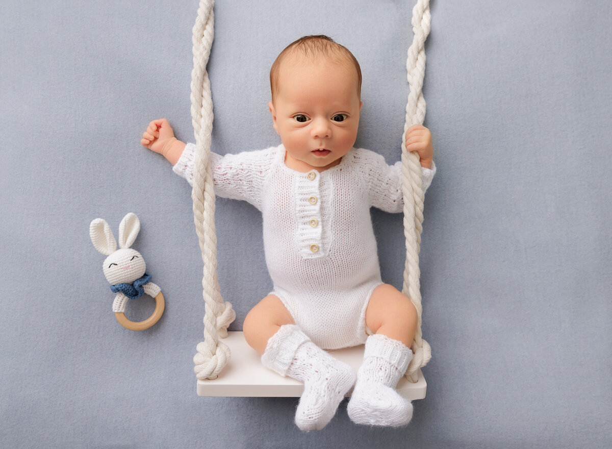Baby boy is awake and alert for his brooklyn ny newborn photoshoot. Baby is in a white knit onesie and booties. There is a swing place under him to give the illusion that he is on a swing. Captured by top Brooklyn newborn photographer Chaya Bornstein