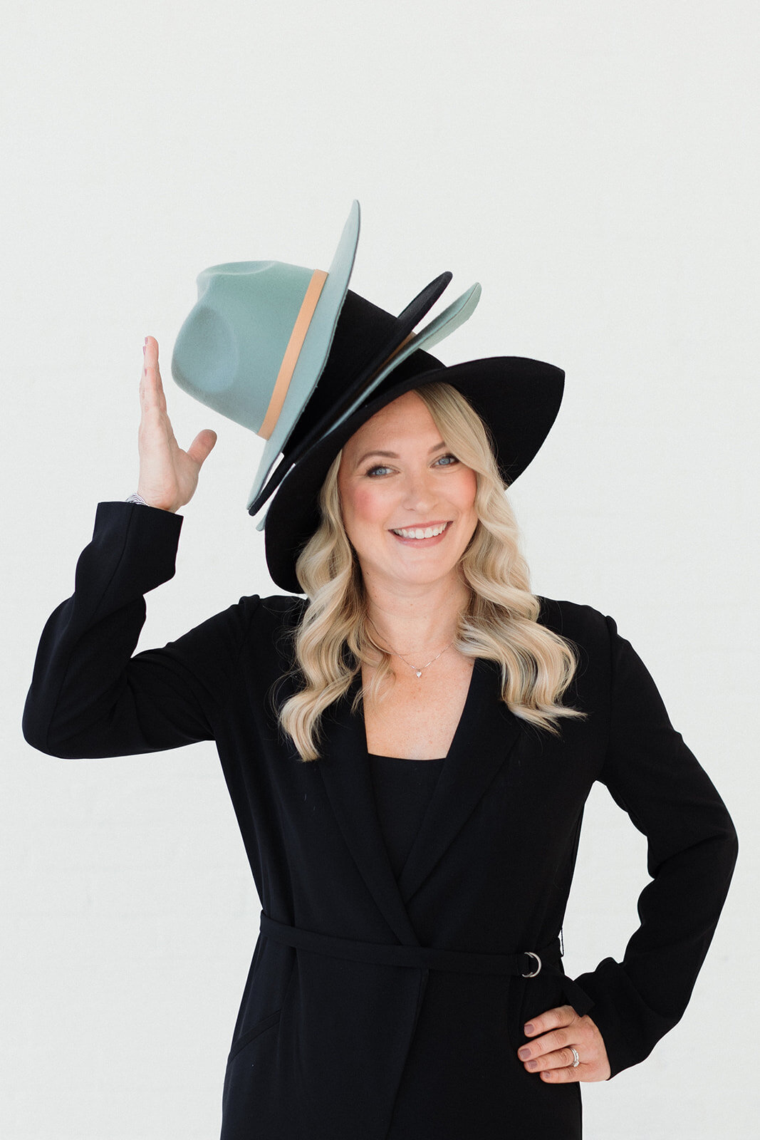 blonde woman has a pile of hats on her head and she is trying to keep them from falling off. She wears a black suit in front of a white backdrop.