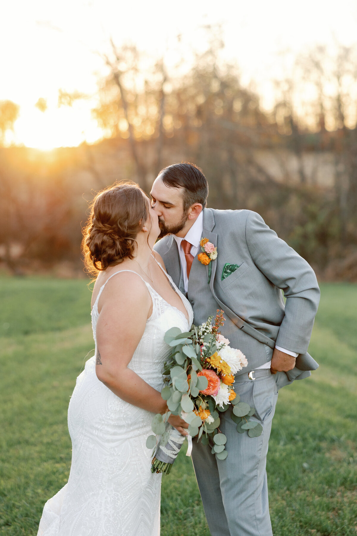 Couple kissing at sunset in an open field at The Hummingbird Chateau, bride in lace gown and groom in light gray suit holding bouquet of white and orange flowers.