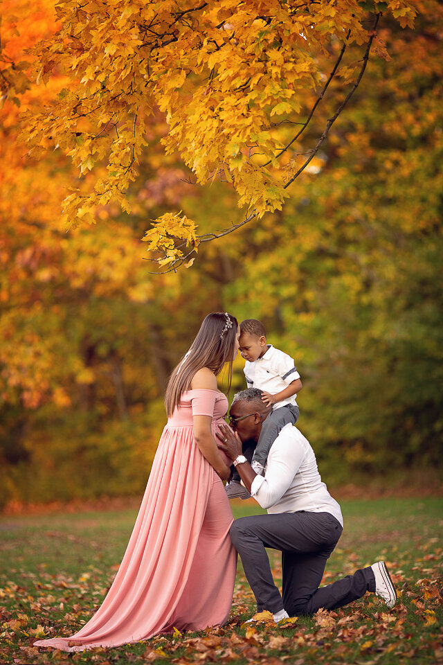 A father kneels in a park to kiss the bump of his pregnant wife with their toddler son on his shoulders