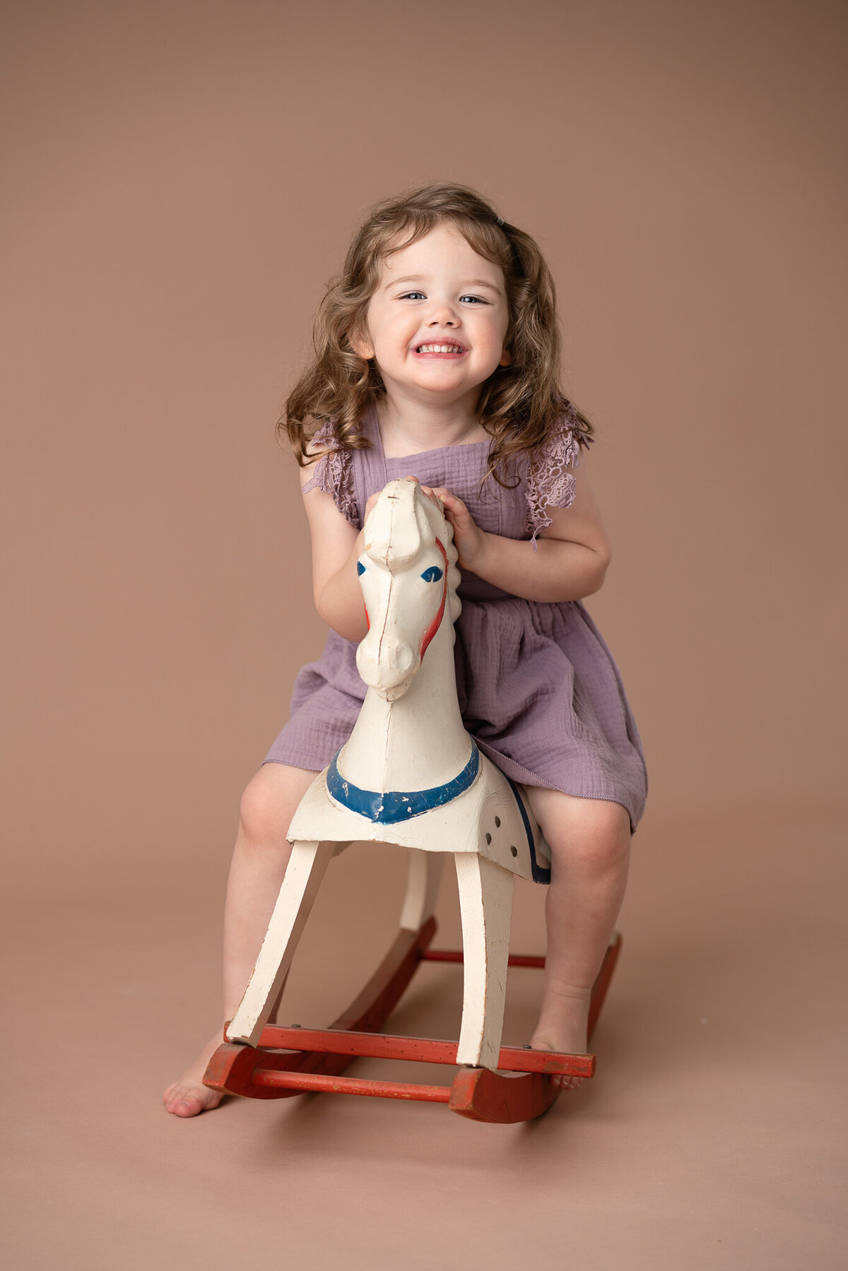 A young girl in a purple dress is riding an old rocking horse and smiling.