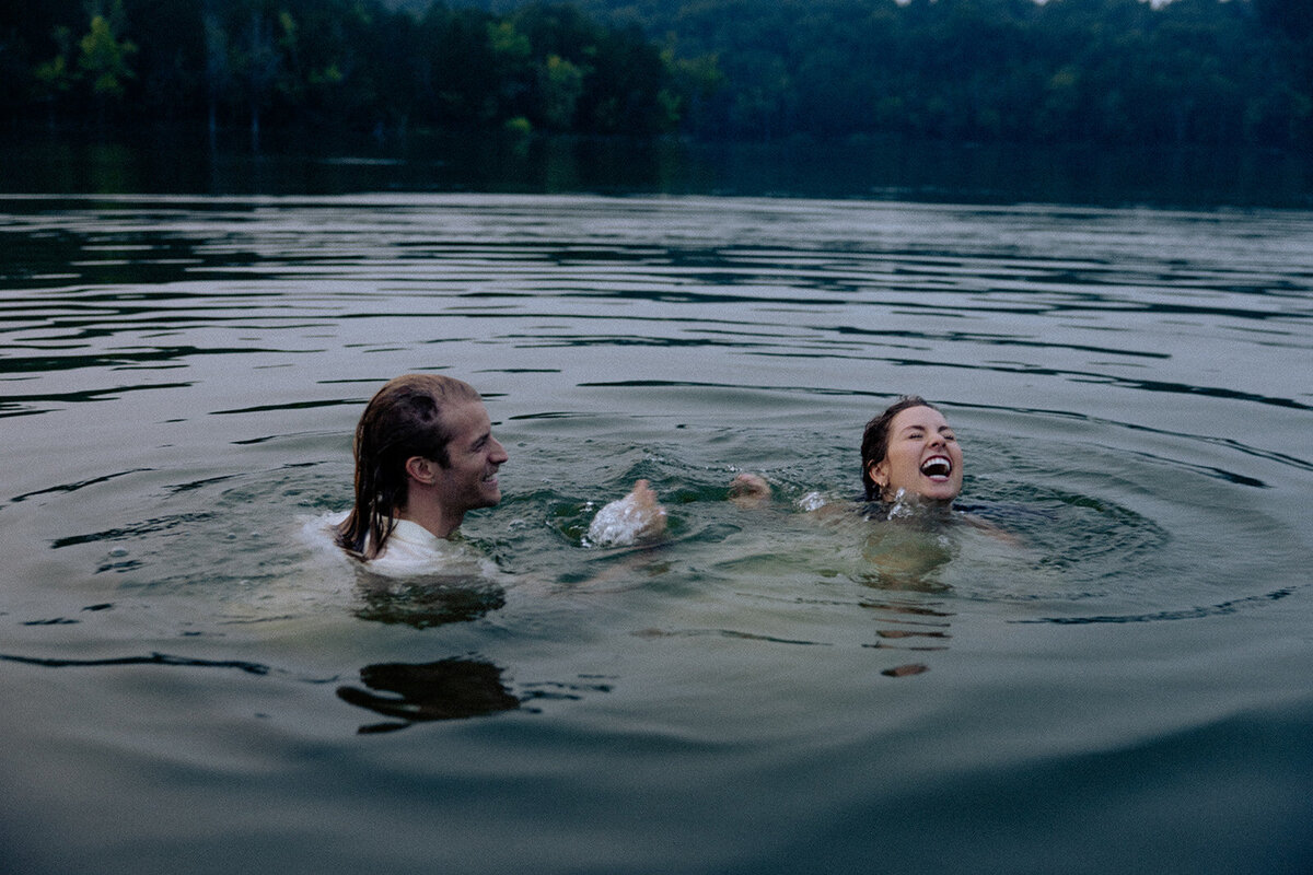 Couple playfully splashes each other while laughing as they have just jumped in a lake during their engagement photo session.