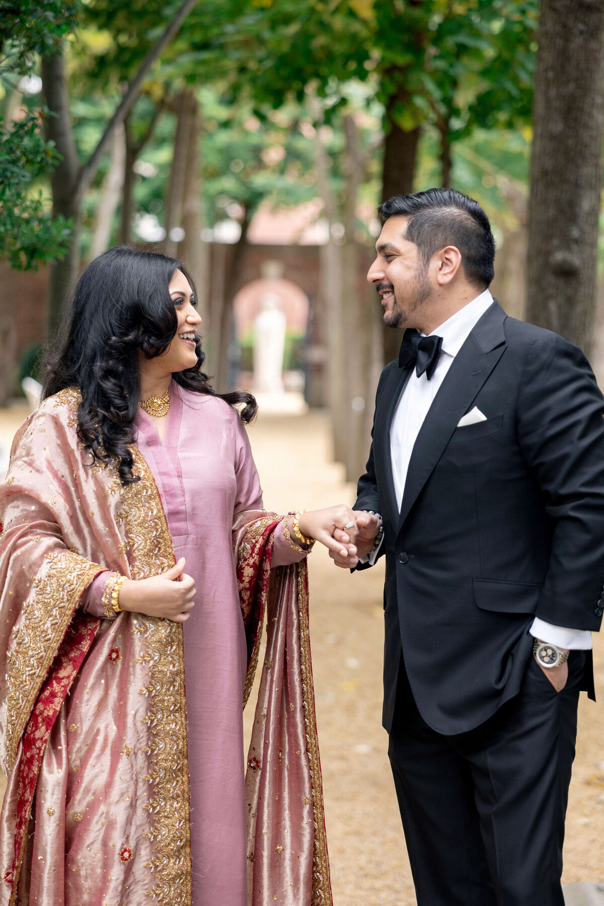 A couple walking hand in hand, smiling at each other. The woman is wearing a pink outfit with a detailed shawl, and the man is in a black suit with a bow tie. They are outdoors on a tree-lined path.