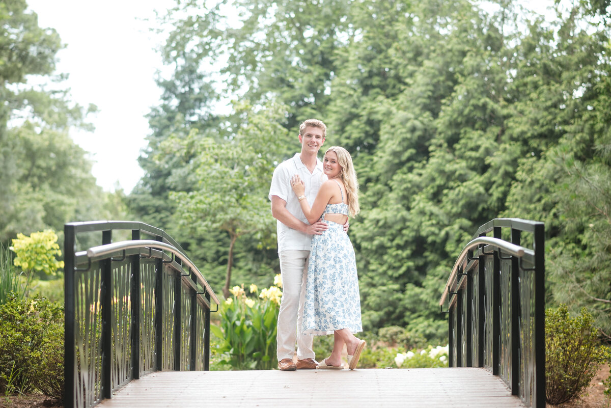 An engaged couple snuggled, smiling, and looking at the camera, standing on the bridge in the WRAL Azalea Garden, surrounded by greenery in Raleigh, NC—captured by DeLong Photography, a top Raleigh wedding photographer, highlighting the most romantic and natural photo locations in the Triangle.