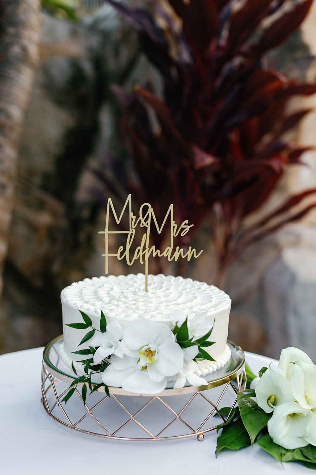 A beautifully decorated white wedding cake with the couple’s last name topper, perfect for a beachside celebration.