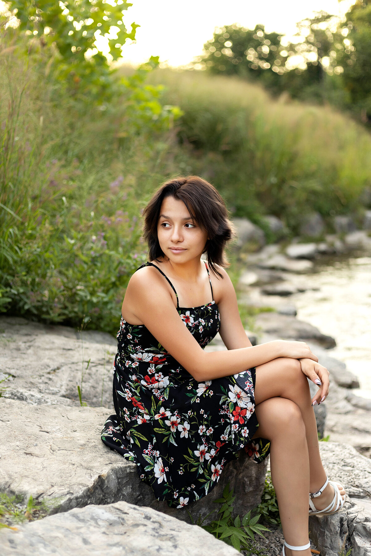 Ann Arbor Argo Park Canoe & Kayak Senior Photoshoot | Ann Arbor Michigan Senior Photographer, Senior girl with black hair in black floral dress sitting on rocks by the Huron River at sunset
