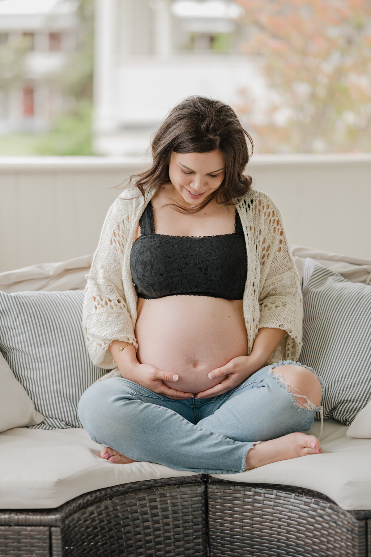 Expecting mom in jeans and an open cardigan sits cross-legged and gazes down at her belly.