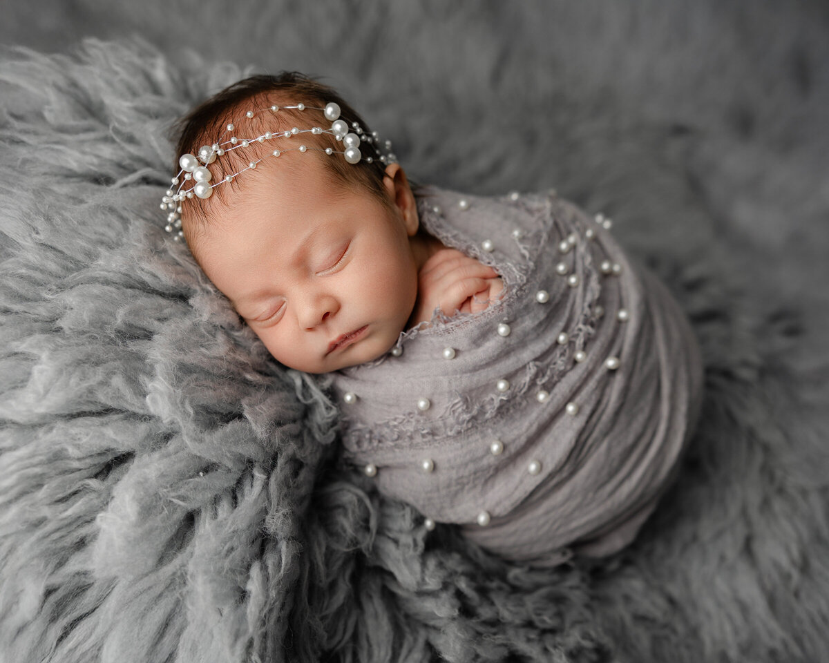 Newborn baby girl swaddled in soft gray fabric, adorned with delicate pearls and a matching pearl headband, posed on a fluffy gray backdrop. Captured by Inland Empire newborn photographer Kaitlyn Dawn Photography