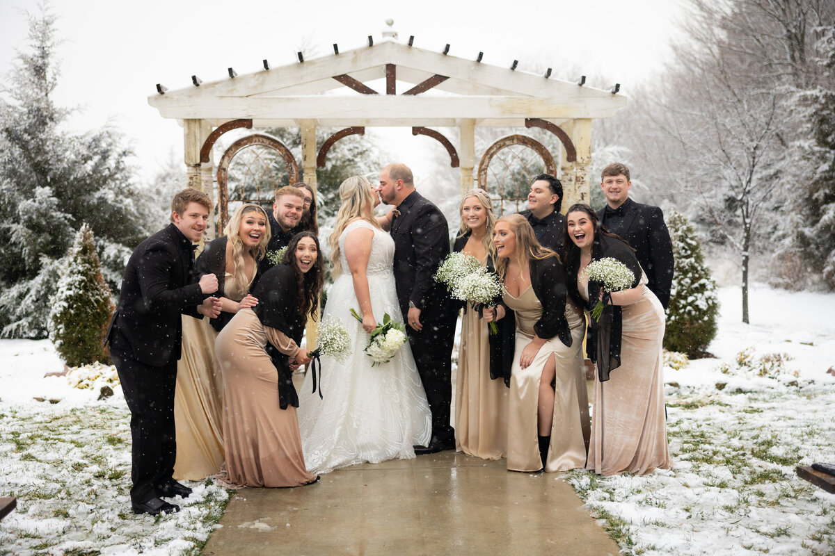 laughing bridal party surrounding the bride and groom while they kiss
