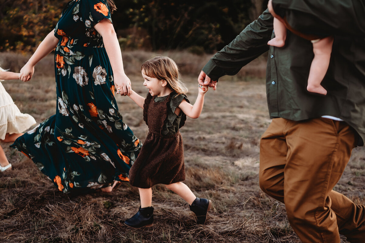 A little girls laughs as she holds her parents' hands and runs in a grassy field, San Diego lifestyle family photographer.