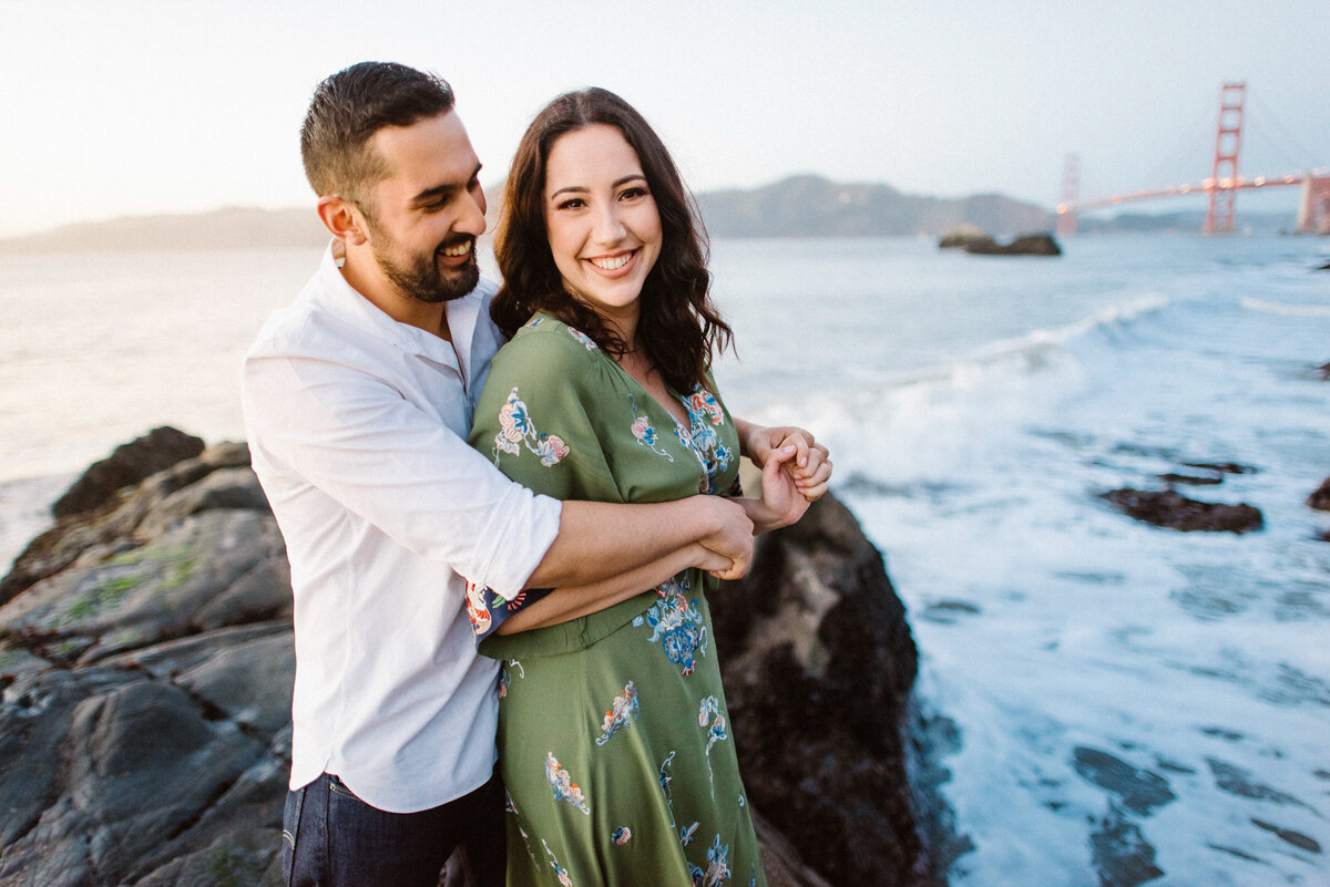 San Francisco engagement photography at Baker Beach with Golden Gate Bridge backdrop. Sunset couples portrait featuring natural candid laughter. Coastal California engagement session with iconic landmark view.