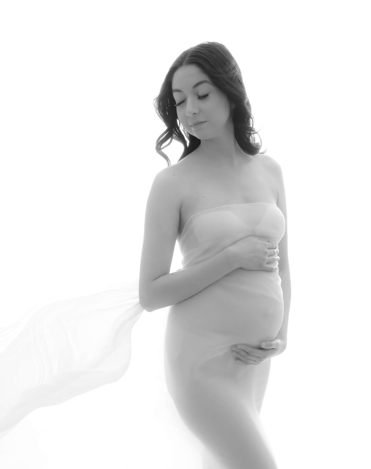 Kitsap maternity session black and white image photographed by Beth Benecki. Mom-to-be is wrapped in a sheer nude color fabric that is tossed by a fan.  Image is cropped and mother is turned slightly to the side and looking over her shoulder.