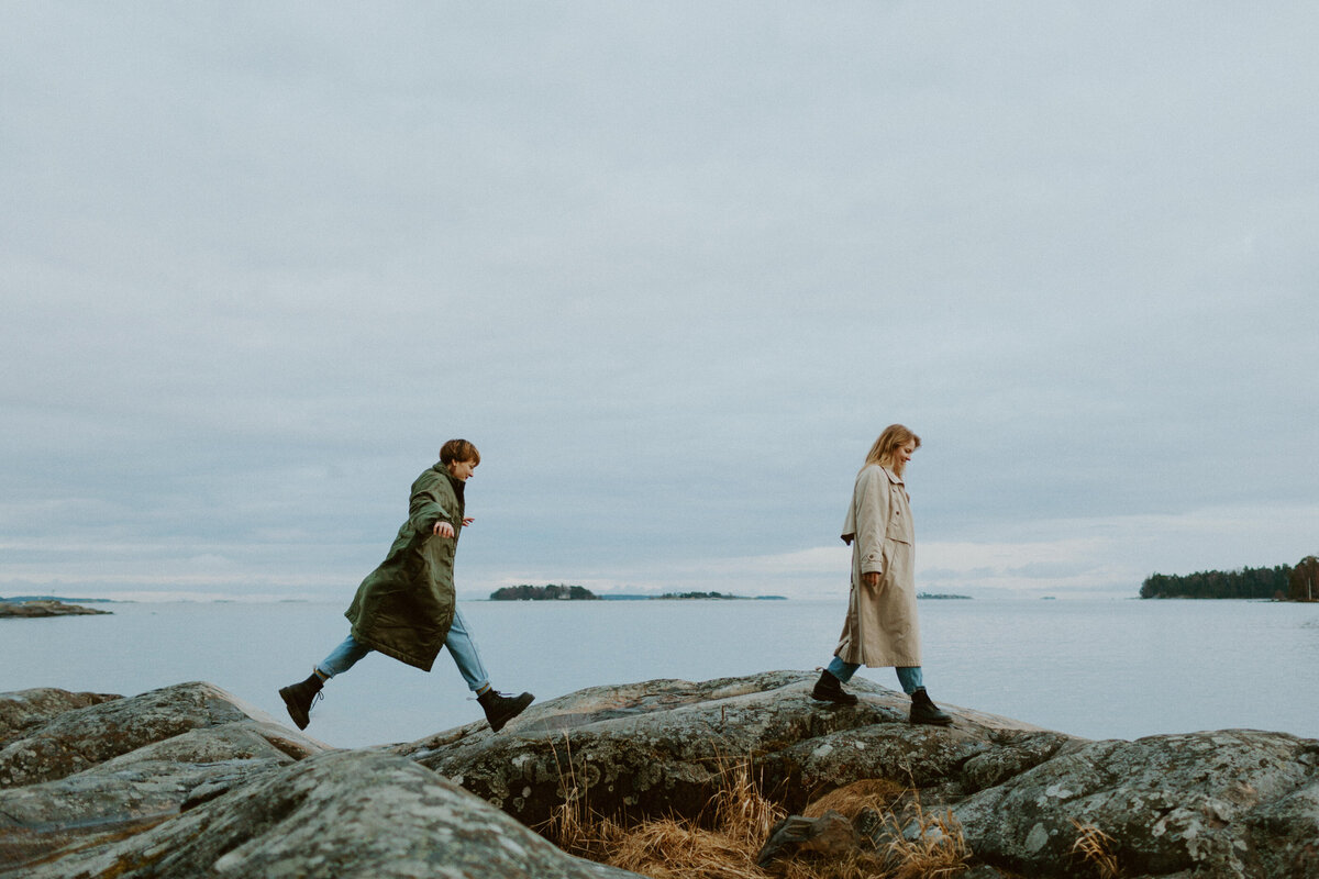 Aikuiset siskot kävelevät rantakalliolla peräkanaa ja takana tuleva hyppää juuri kolon yli Kallahdessa Helsingissä.
