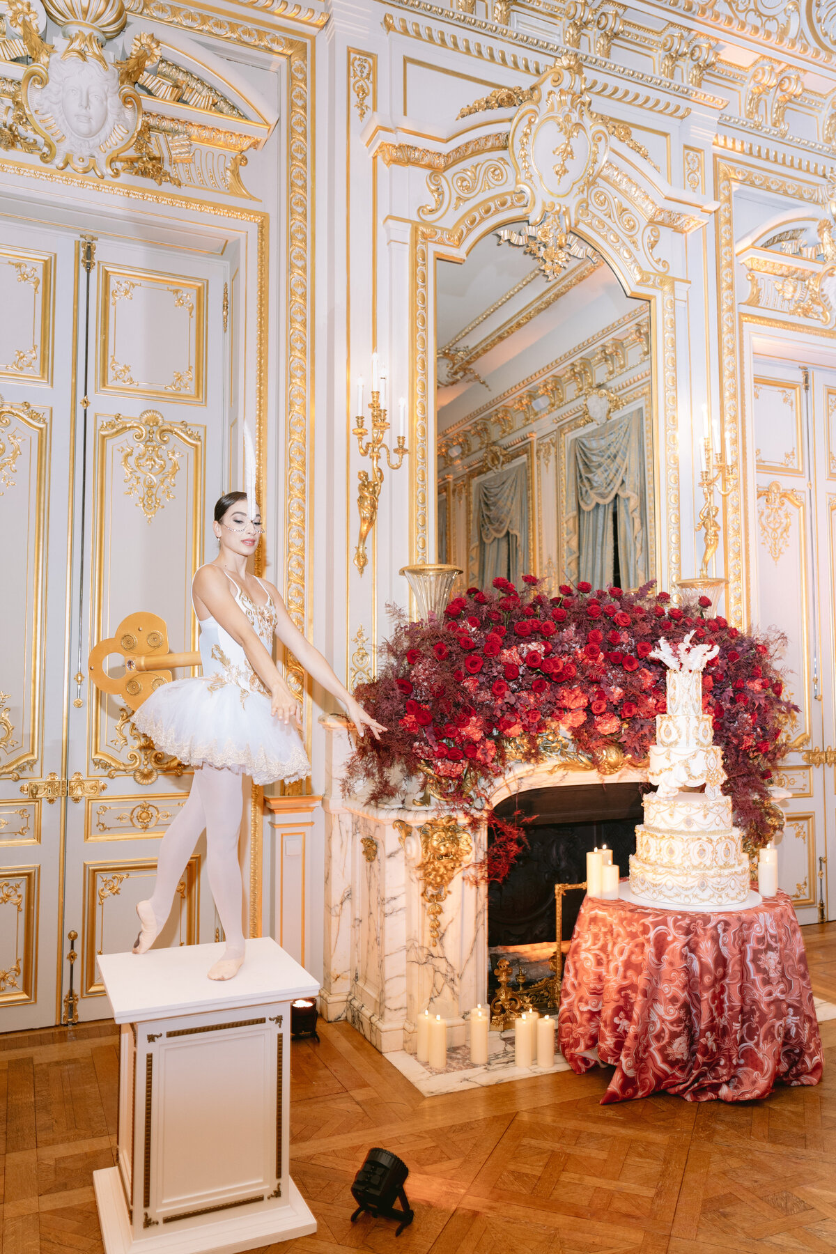 The photo was taken at Shangri La Hotel & Ritz Paris Hotel in Paris, France by the talented photographer Matteo Coltro, with Zen Film Works as Videographers. In the luxurious setting, a ballet dancer in a white tutu poses elegantly atop a pedestal. She is gracefully positioned beside a fireplace adorned with a large bouquet of red flowers and a tall, ornate white cake. The room itself is a sight to behold with its gold-trimmed walls, intricate moldings, and elegantly draped curtains, creating a truly opulent backdrop for the scene.