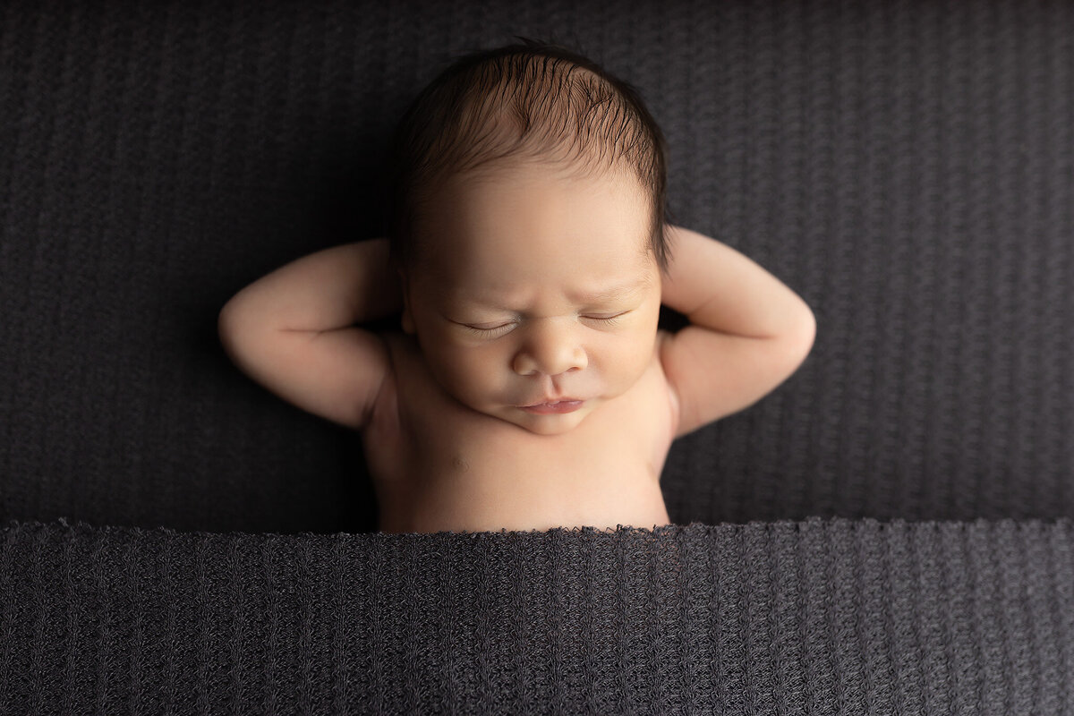 Newborn tucked under gray blanket, hands behind head — baby photographer newborn session.