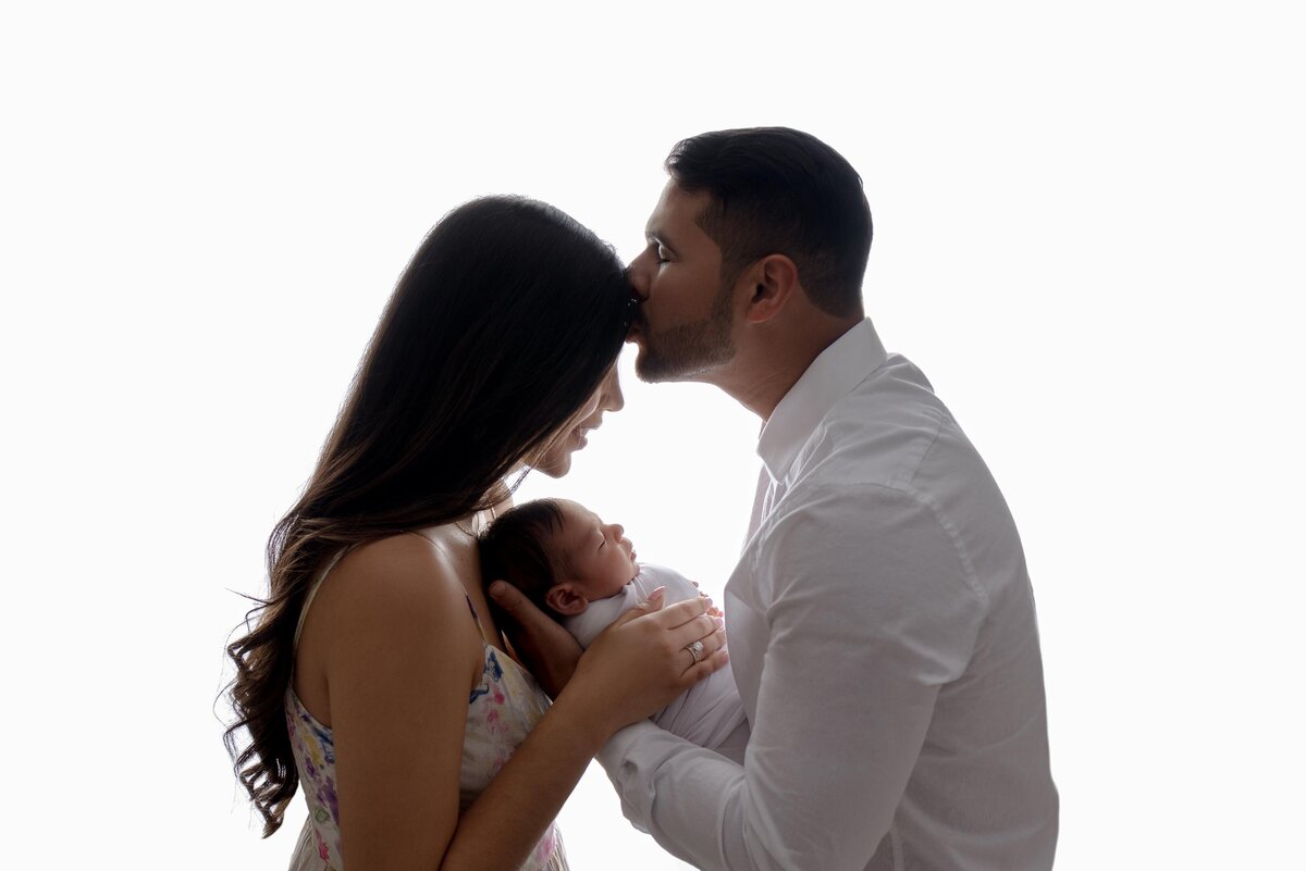 Profile backlit image of new parents holding baby with dad kissing mom's forehead