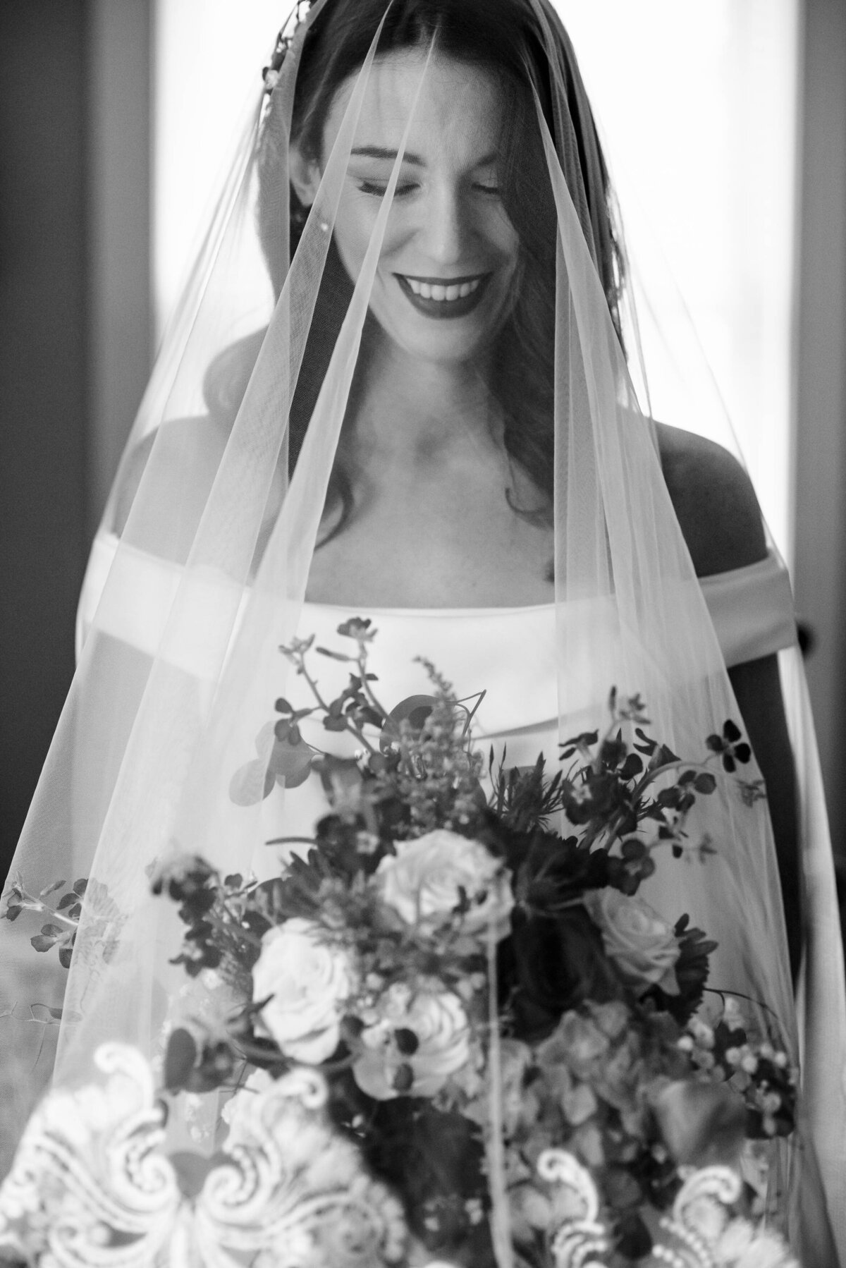 Black and white portrait of a bride smiling and looking at her bouquet with her veil over her face  by Charlotte wedding photographers DeLong Photography
