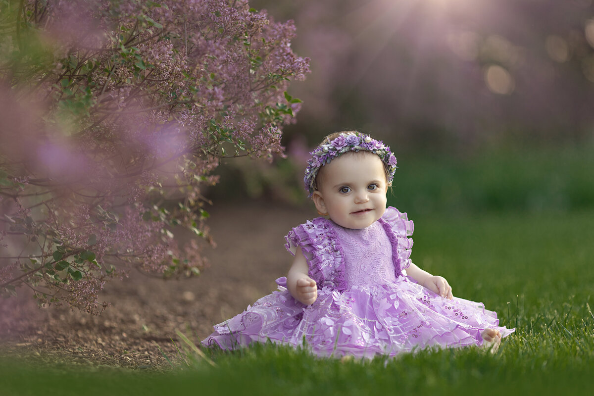 A toddler girl in a purple dress and matching floral headband sits in a grassy lawn at sunset taken by a New Jersey Family Photographer
