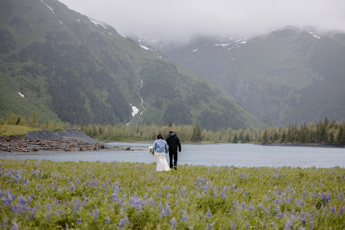 Alaska elopement couple walks towards lake with lupine field behind them and mountains ahead of them.