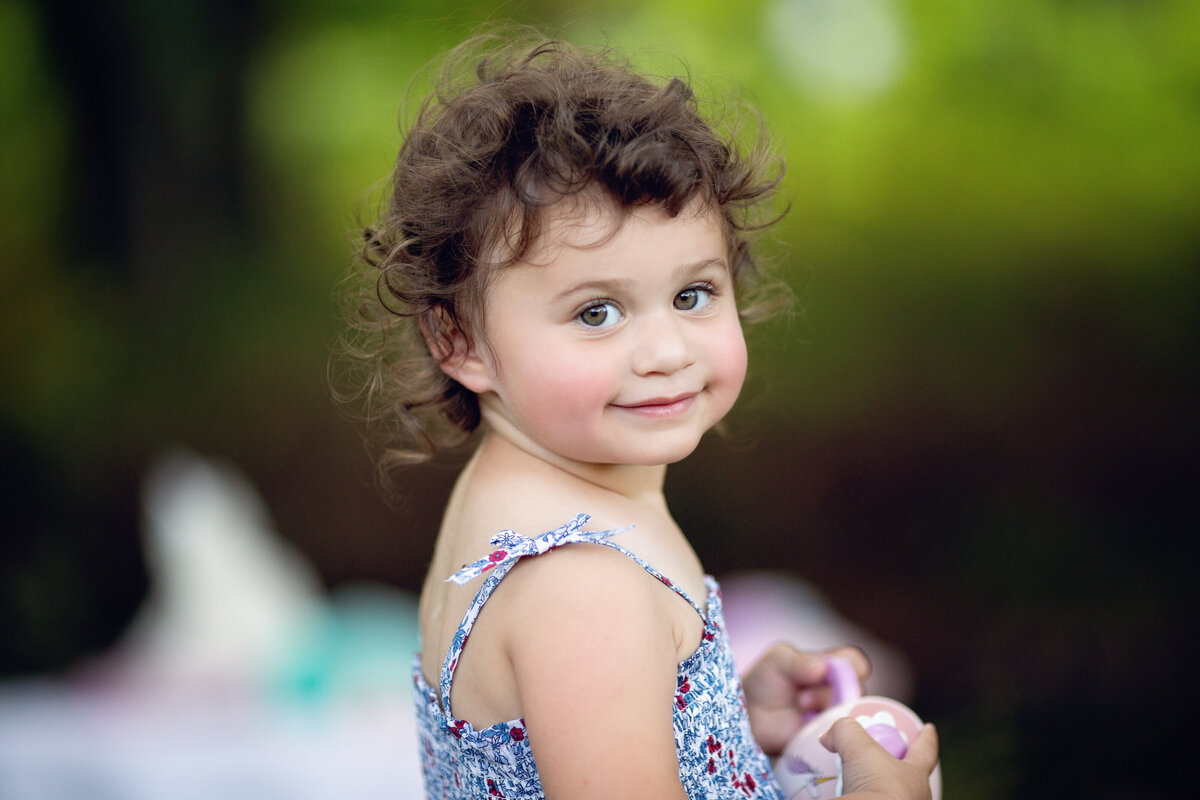A toddler girl smiles over her shoulder while playing with toys in a bathing suit in a park