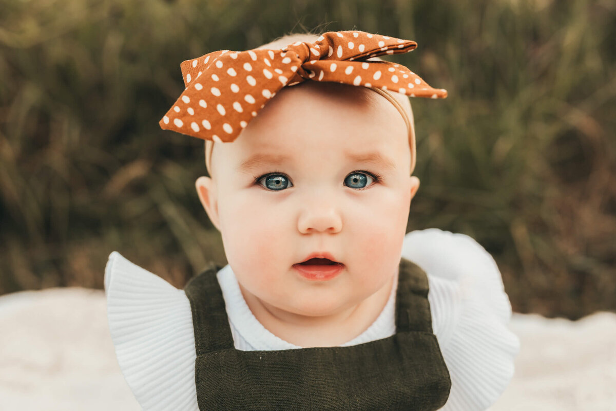 little girl wears an olive romper with white shirt and a rust colored polka dot bow.