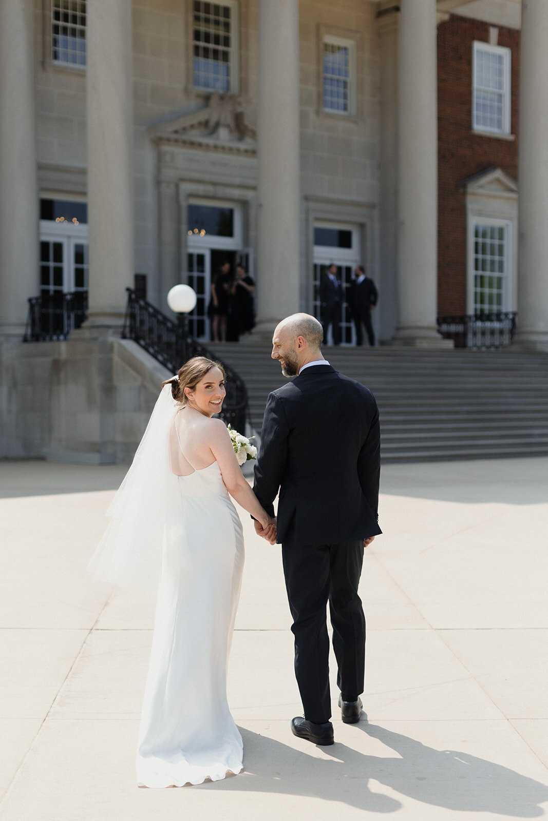Couple walks toward the Chicago History Museum and bride turns to look at camera, as groom looks at her. Groomsmen stand on the steps in the distance.