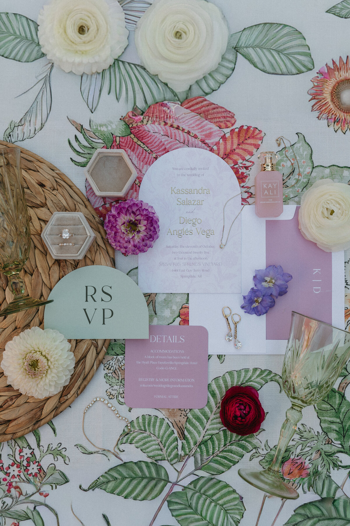 Wedding invitation suite flatlay featuring pastel flowers, vintage glassware, jewelry, and textured linens, showcasing elegant stationery styling with soft floral details.

