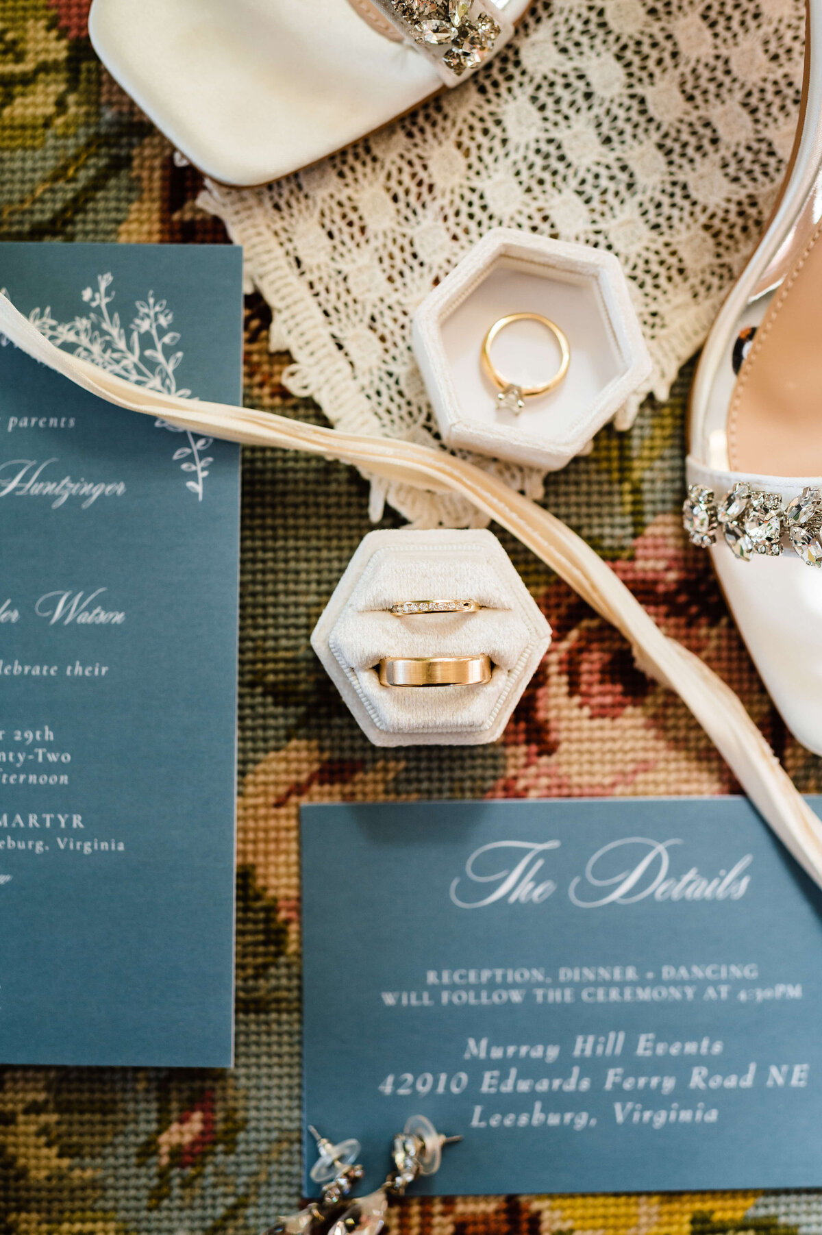 bride and grooms wedding rings sitting in a a white velvet box next to the wedding invite and save the date on a vintage tapestry with green and blue under tones