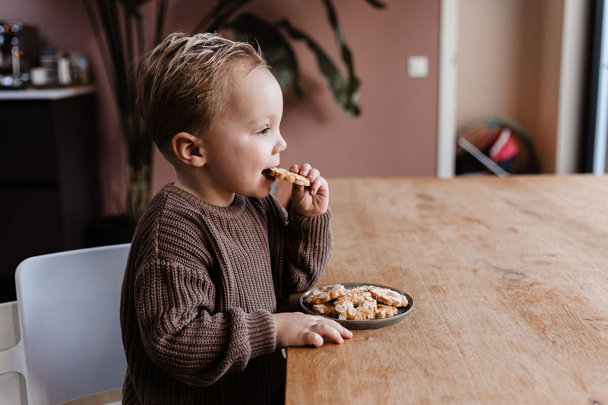 Jongetje zit aan tafel met schaal koekjes voor zich en eet een koekje met lach op het gezicht
