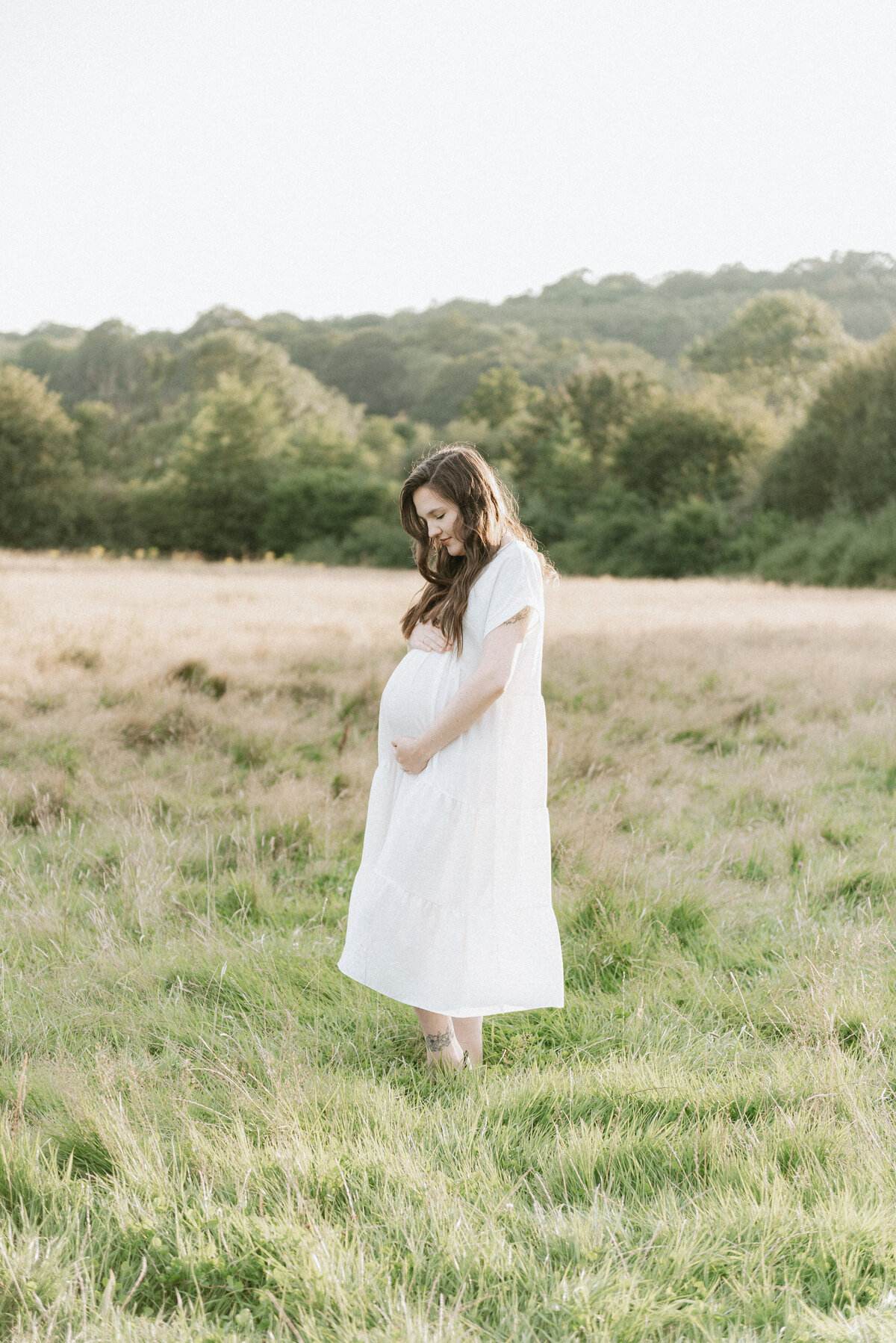 A women standing in a meadow in a white dress holding her pregnant bump at a photoshoot in Billingshurst