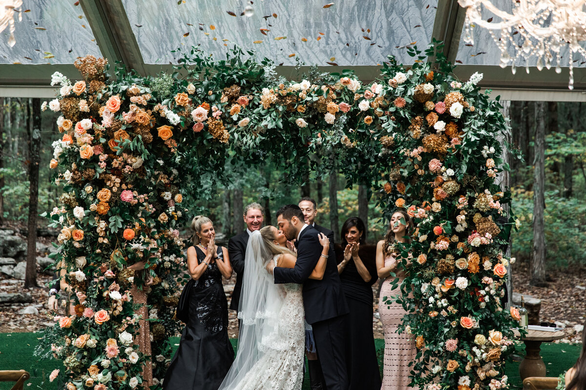 Eye-catching oversized chuppah overflowing with fall florals featuring dahlias, garden roses, rain tree pods, and fall greenery. Autumnal hues of terra cotta, dusty pink, copper, and yellow create this statement wedding piece. Designed by Rosemary and Finch in Nashville, TN.