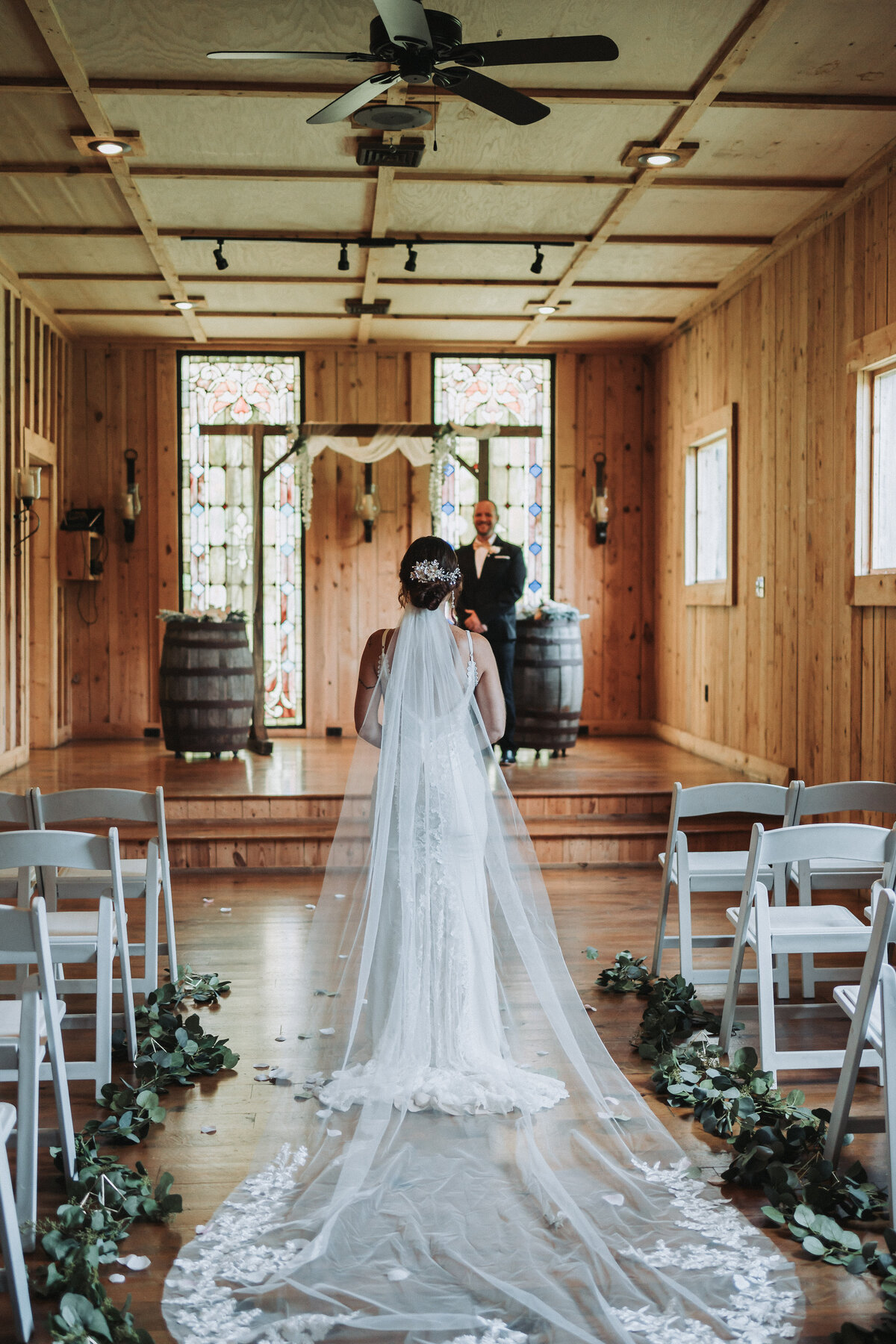 Bride walks down a greenery-lined aisle toward her groom for a first look inside the stained glass chapel at The Gentry Farm in Danville, Virginia.