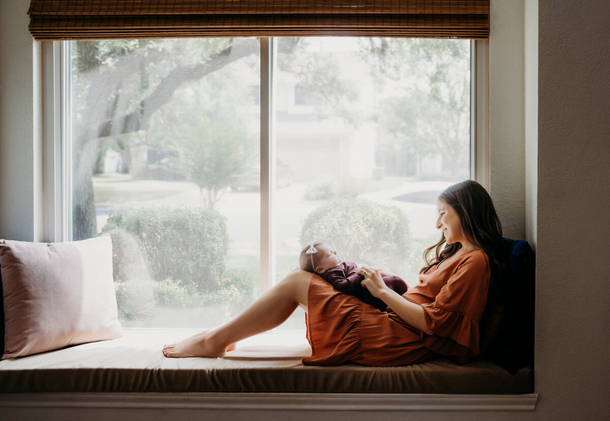 Newborn Photographer, a woman sits in a window nookholding her baby