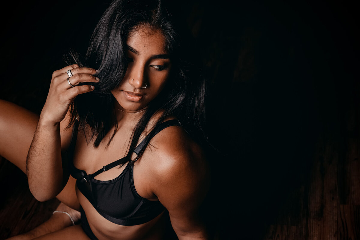 An image from a DC boudoir photography studio of a woman in a black bra gazing over her shoulder in a dark room with a hand in her hair