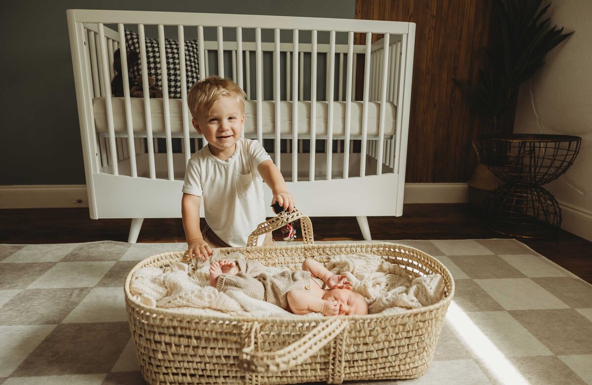 Young kid is watching  his new baby sibling who is sleeping in a basket on the floor of a bedroom