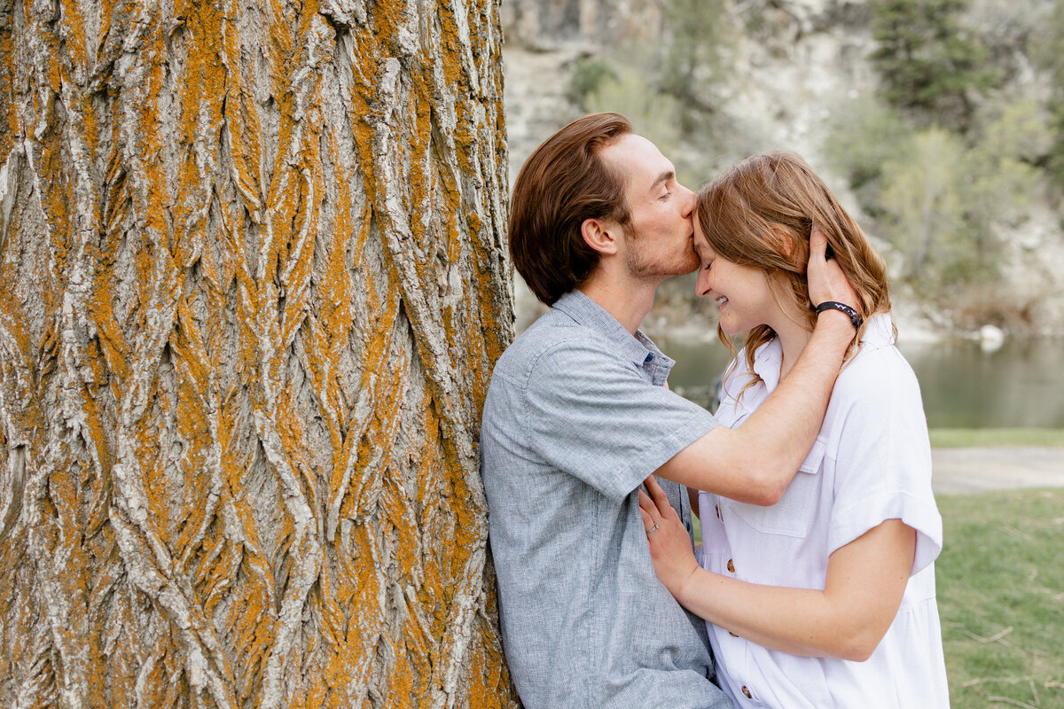 A man kisses his fiancée’s forehead while she smiles, leaning against a rustic tree in the forested landscape of Provo Canyon.