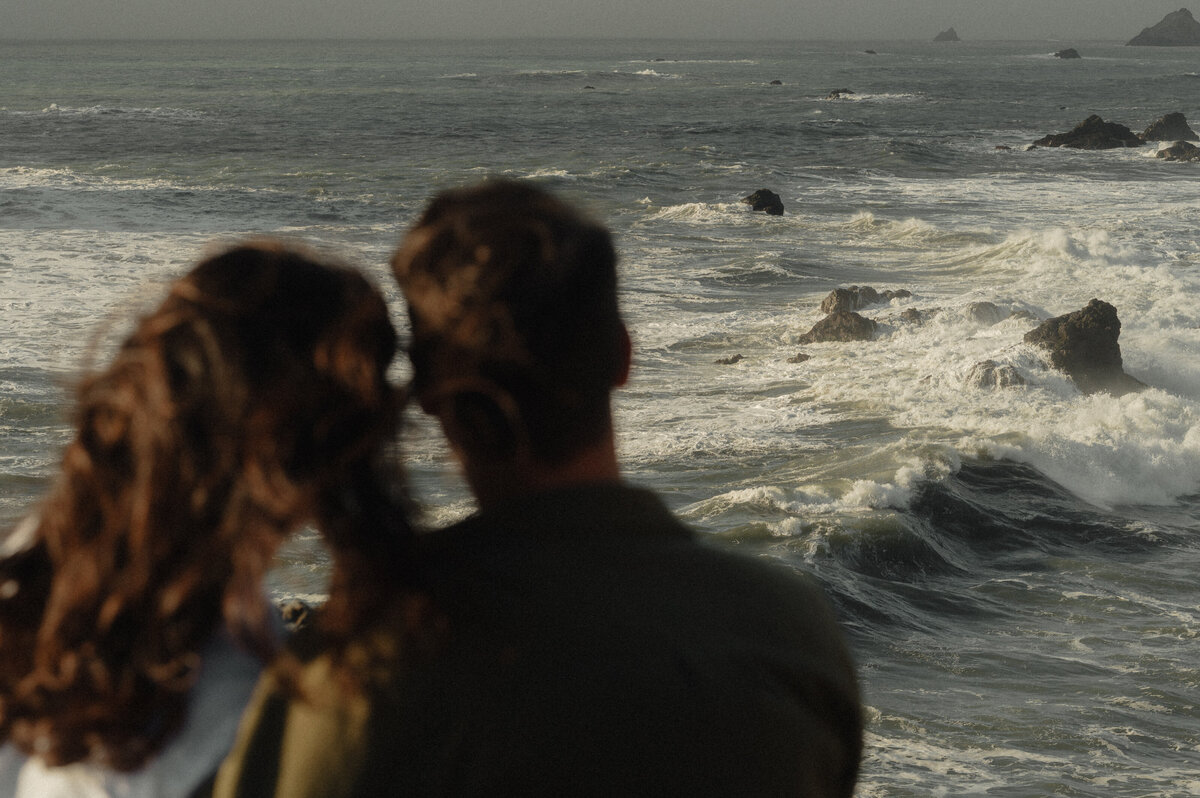 windy Oregon beach engagement photo with couple cuddled close and looking out at the ocean
