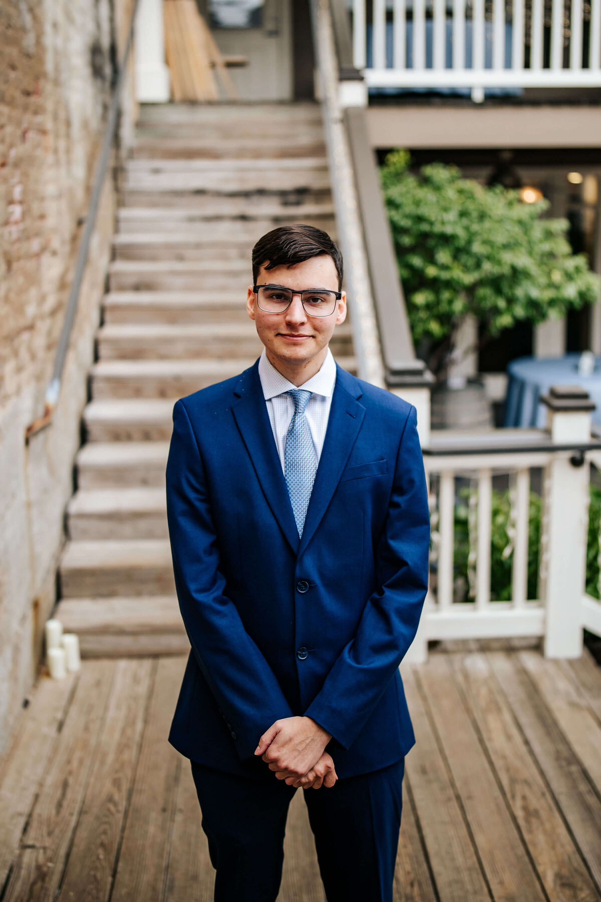 A groom in a blue suit and tie stands with hands clasped in front of him on a wooden deck, with stairs and greenery in the background.