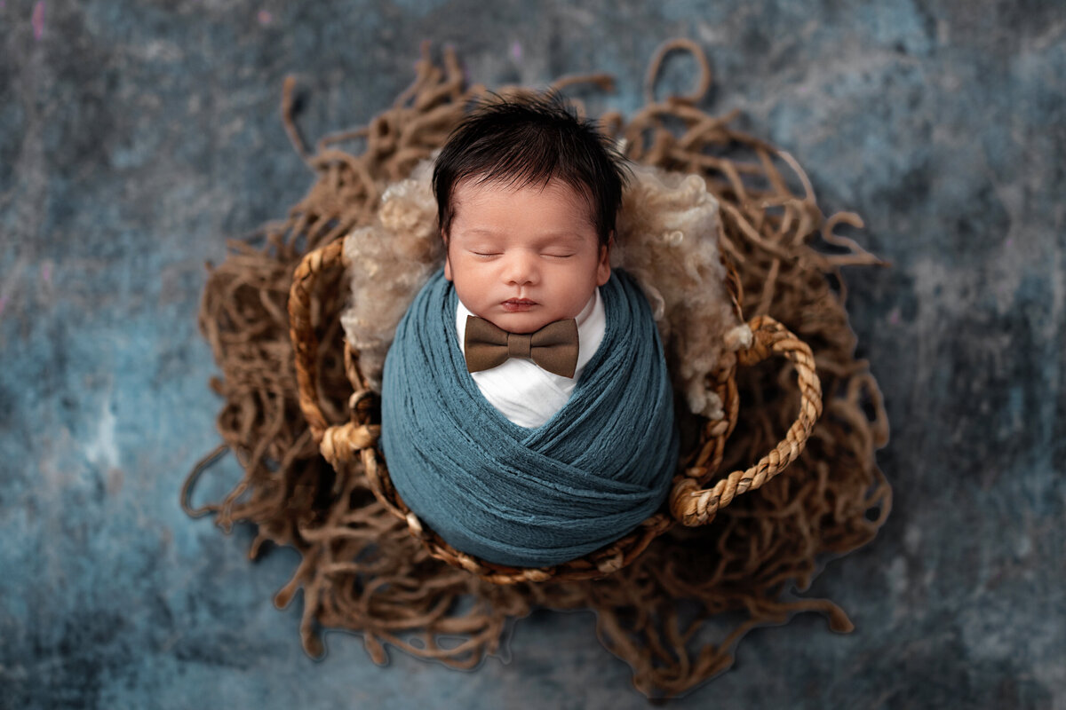 Newborn baby boy with bow tie wrapped in deep blue inside a rustic wooden bowl on textured blue background.