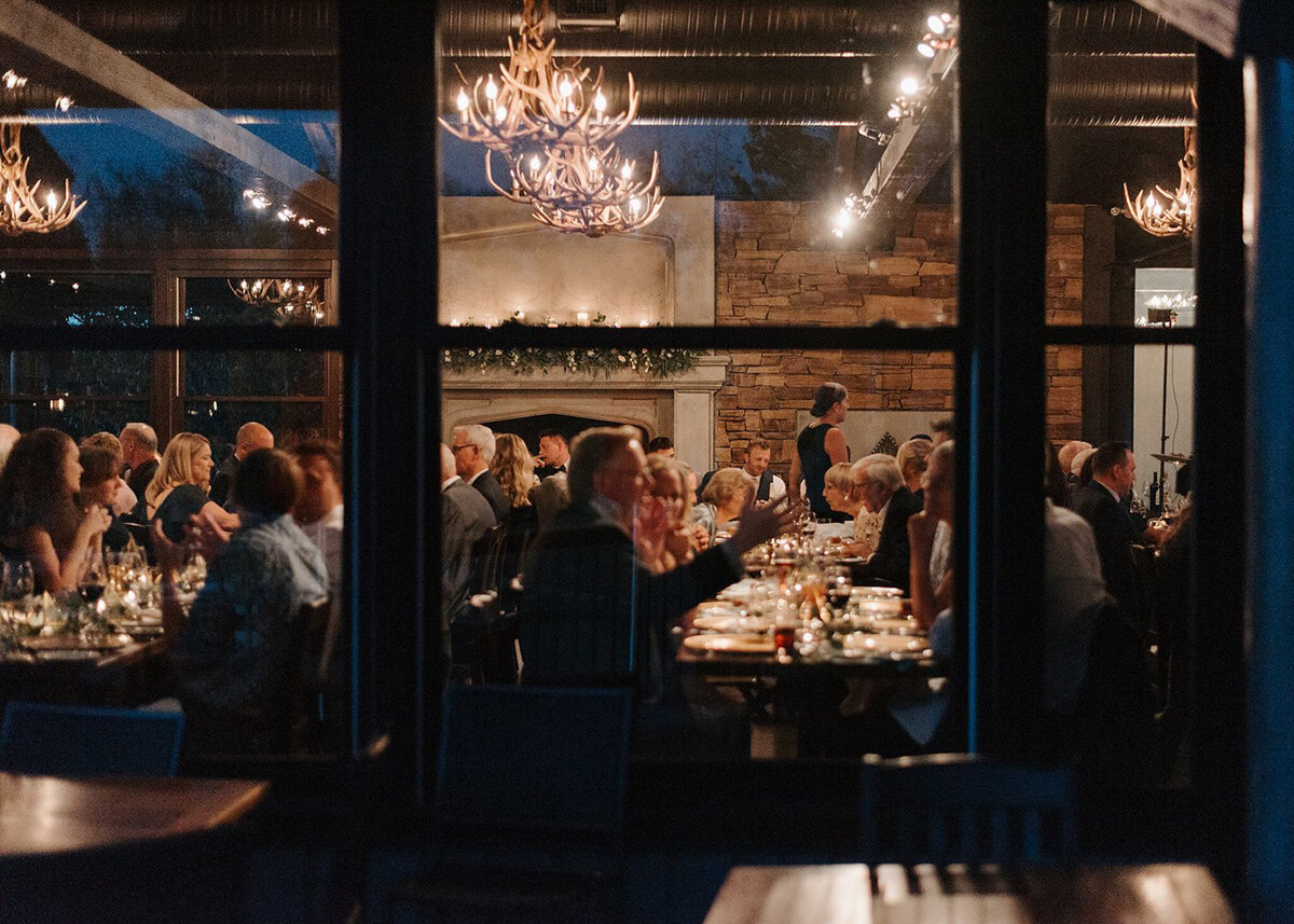 A warm and intimate wedding reception at The Lake House in Calgary, viewed from outside through large glass windows. Guests are seated at long, candlelit tables, engaged in conversation and enjoying the elegant atmosphere. The rustic charm of the venue is highlighted by stone walls, a grand fireplace adorned with greenery and candles, and antler chandeliers casting a soft glow. The dim lighting and reflections in the glass add depth to the scene, creating a cozy and romantic ambiance for the celebration.