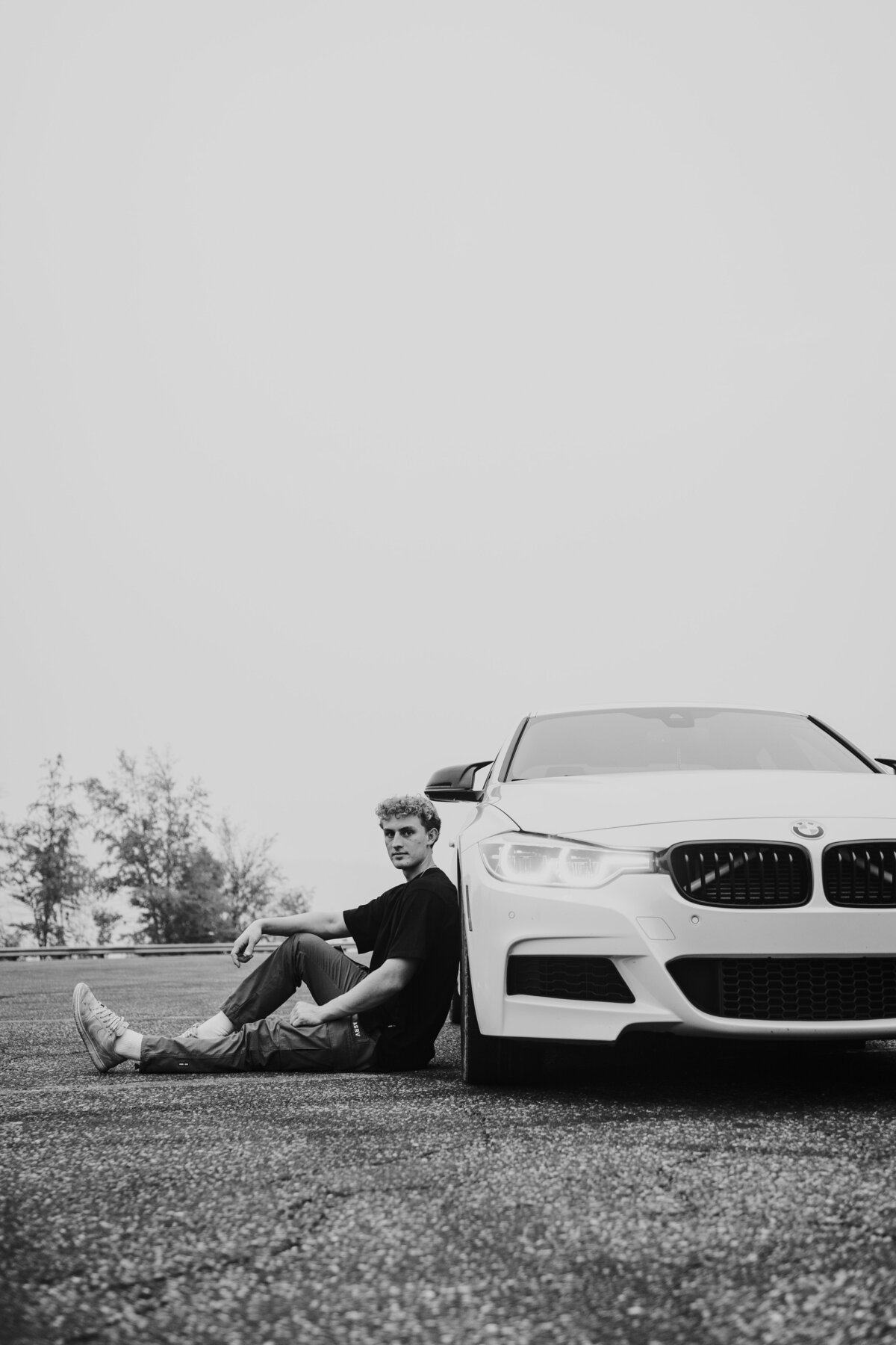 Black and white shot of young man leaning against the side of white BMW in a parking lot.