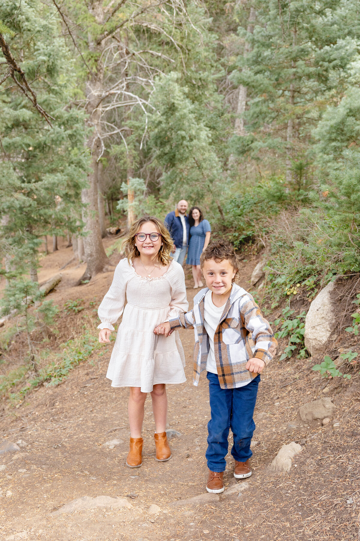 A young boy and girl joyfully run along a dirt trail in the forest, their parents standing behind them, slightly out of focus. The setting is a beautiful, lush mountain landscape.