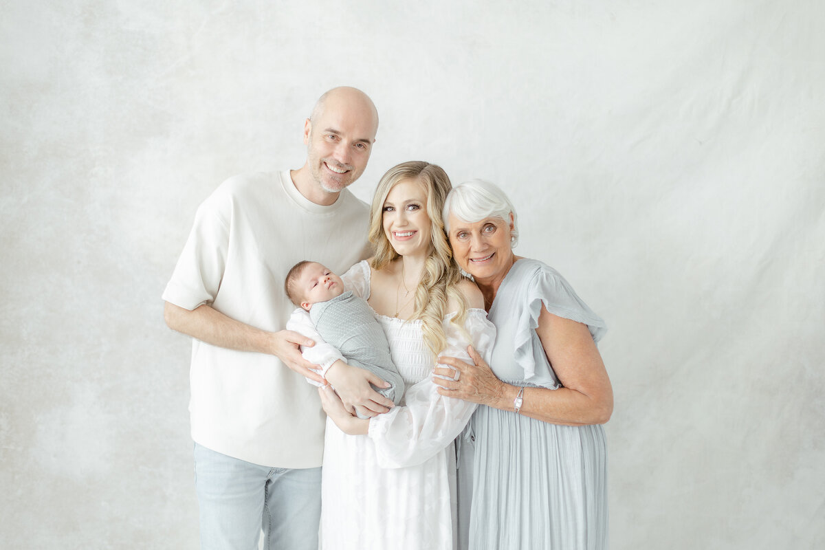 An in studio family portrait of mom, dad, their newborn baby and the grandmother as they are all snuggled together for their family photos taken at a Dallas/Fort Worth photography studio.